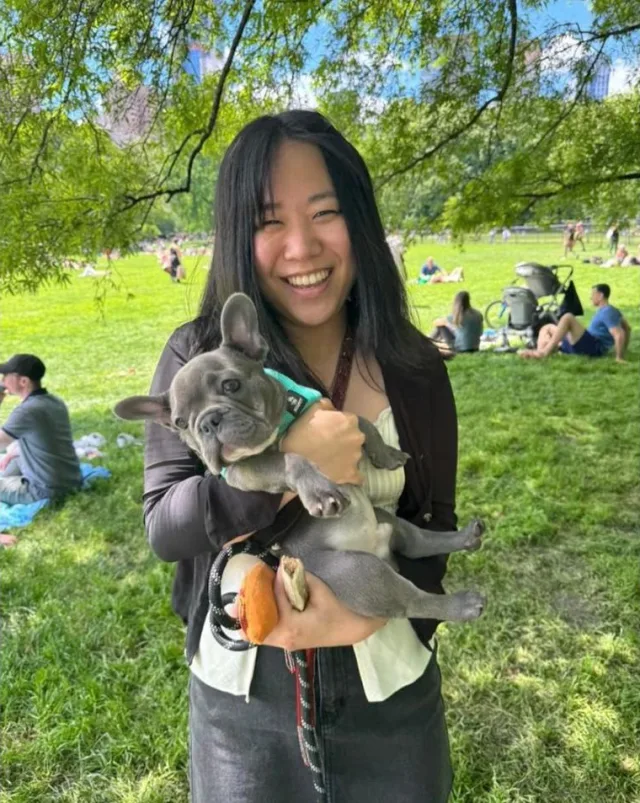 Smiling woman in a park holding a grey-blue Frenchie puppy in a teal harness